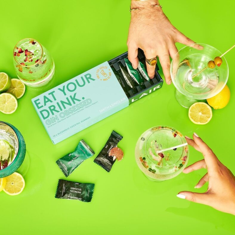 An image of a Smith & Sinclair gin cocktail gummies box alongside some hands and glasses of gin